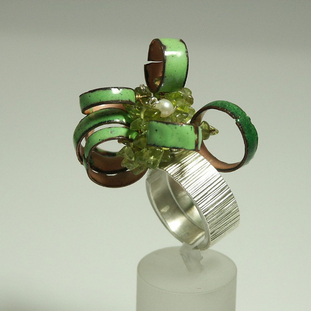 Bague décorative unique avec des bandes en métal vert et des pierres précieuses ornées, présentée sur un support.