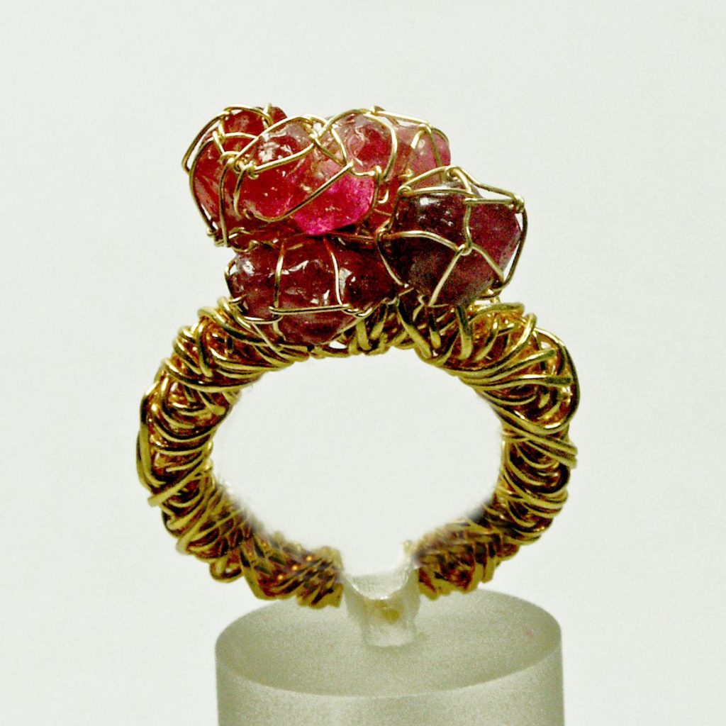 Bague en filigrane doré ornée de pierres précieuses roses, présentée sur un support transparent.