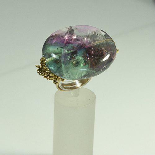 Bague en pierre précieuse multicolore avec des nuances de vert, de violet et de rose, montée sur une base en métal doré.