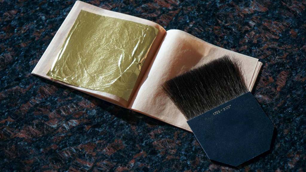 Un livre ouvert avec une feuille de dorure en or et un pinceau sur une surface texturée sombre.