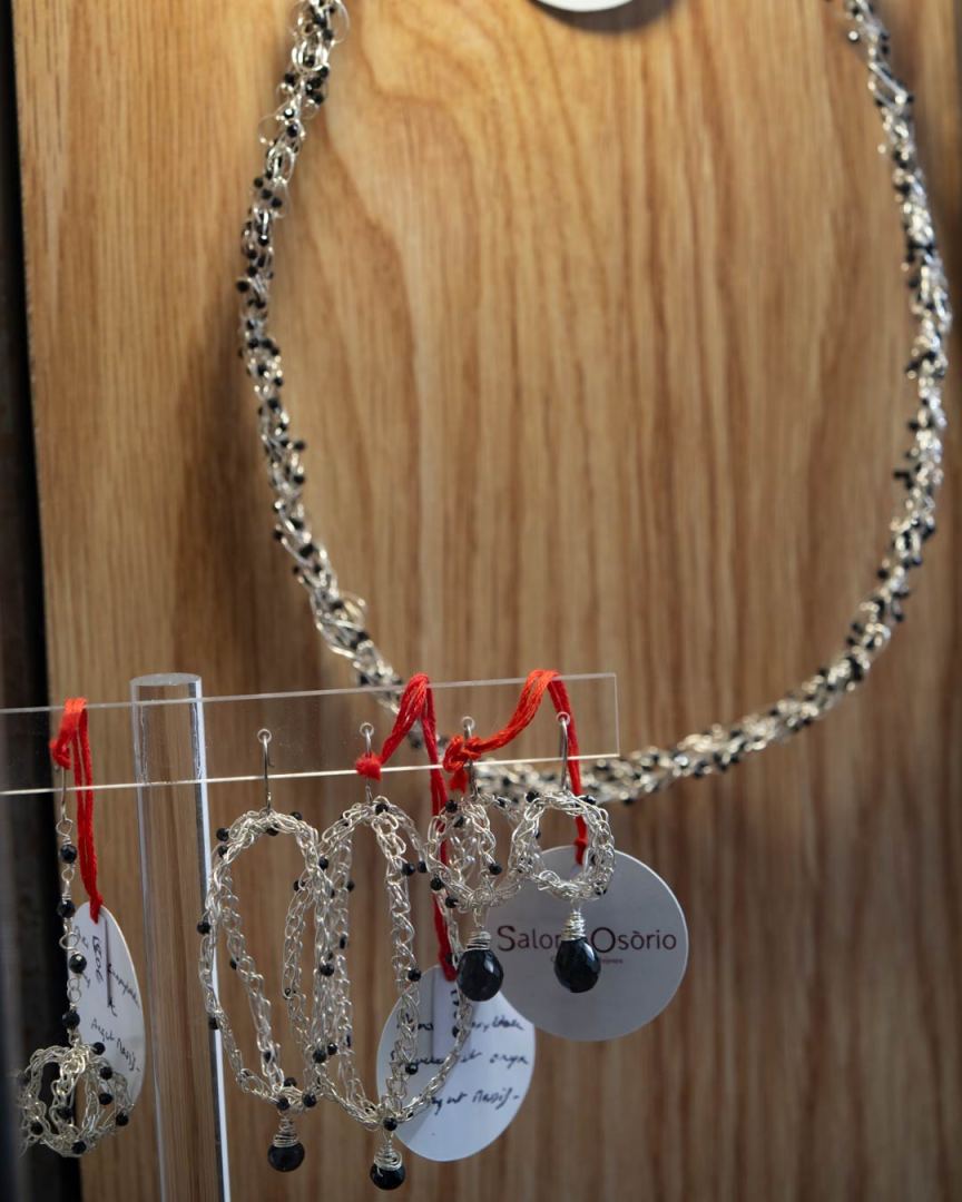 Collier en argent avec des perles noires suspendu sur un fond en bois, accompagné de boucles d'oreilles assorties sur un support transparent.